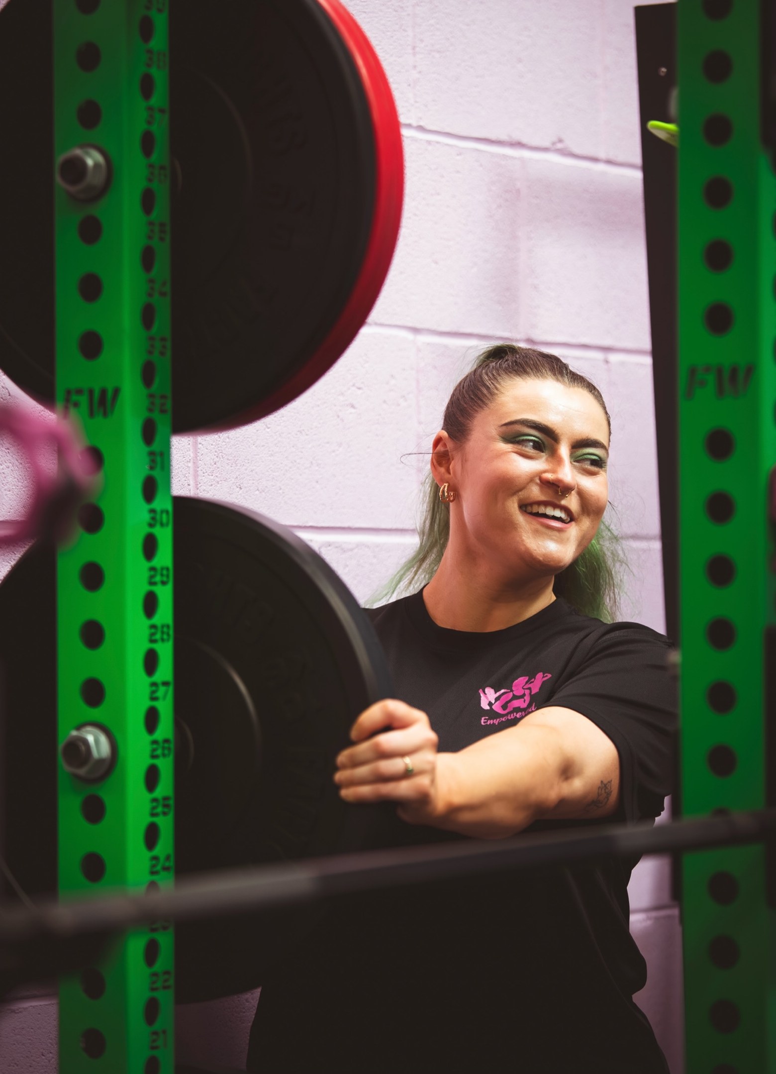 A smiling coach in a black Empowered tee leaning on a green squat rack against a pink wall
